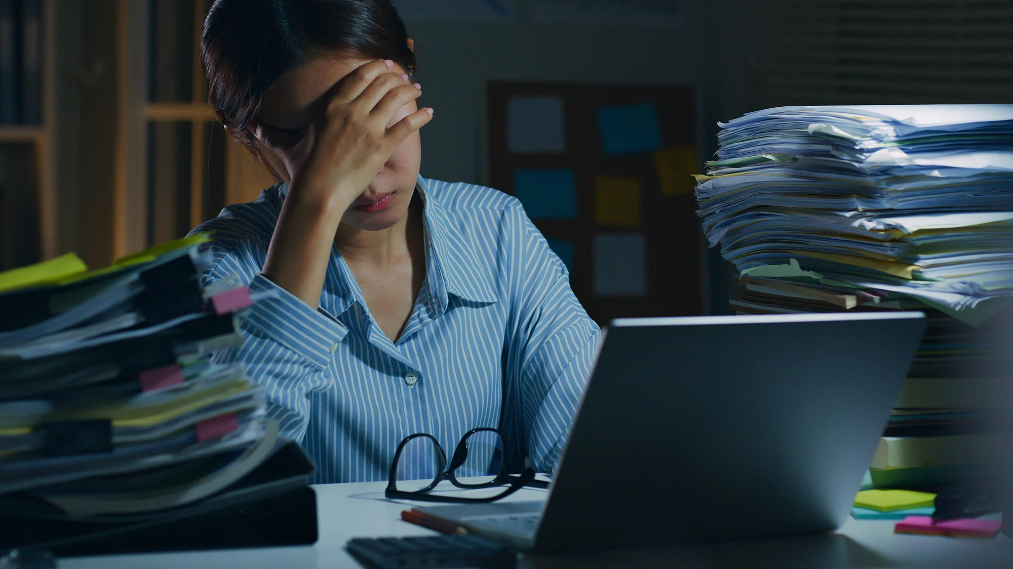 Stressed office worker sitting at a desk with a laptop, surrounded by stacks of paper invoices and business documents.