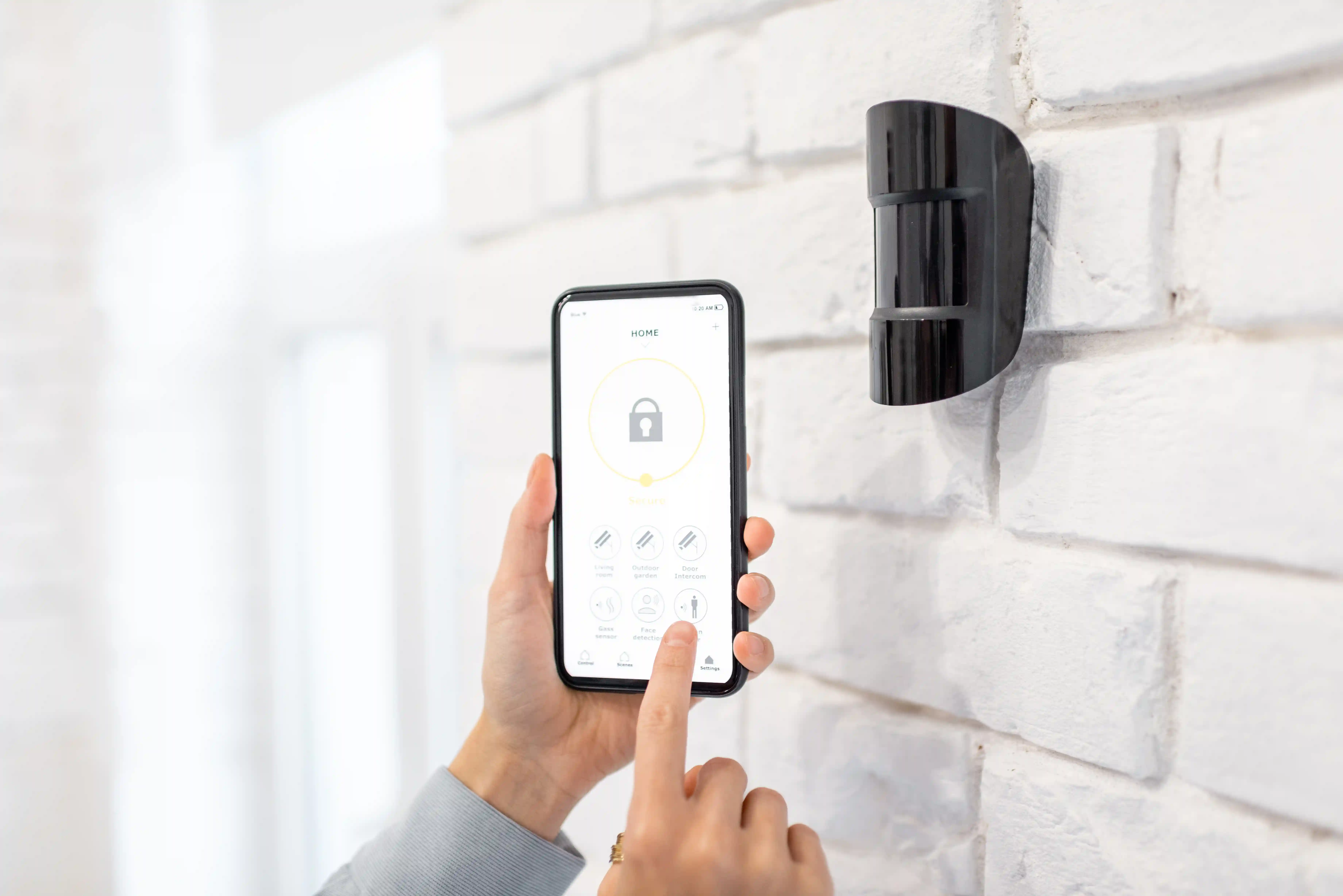 Person using smartphone to control smart home security system with sensor on wall.