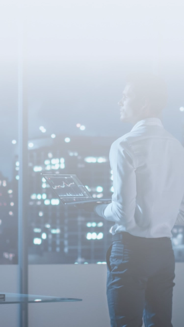 Businessman analyzing data on laptop while overlooking city skyline through office window at night.